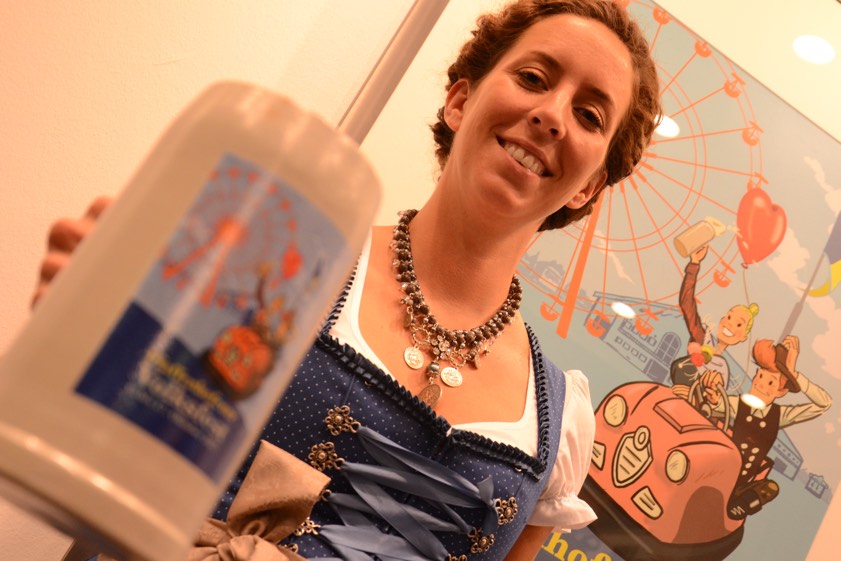 Festwirtin Julia Spitzenberger präsentiert den diesjährigen Volksfest-Krug, den die Stadtverwaltung in limitierter Auflage verkauft. Im Hintergrund das aktuelle Volksfest-Plakat, gestaltet von Ferdinand Haschner. Das Motiv findet sich auch auf dem Humpen wieder. Fotos: Zell